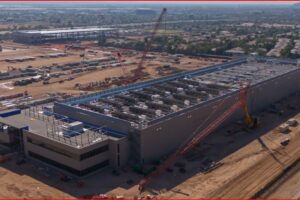 A long industrial building under construction seen from above, with multiple cranes and a dusty work site surrounding the structure.