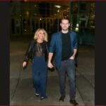 A couple walks hand-in-hand outdoors at night; the woman wears a black leather jacket and navy dress, the man wears a blue blazer and dark jeans.