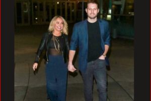 A couple walks hand-in-hand outdoors at night; the woman wears a black leather jacket and navy dress, the man wears a blue blazer and dark jeans.