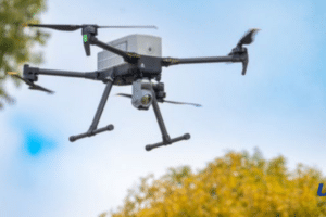 Quadcopter drone with a mounted camera flying over trees, blue sky in the background, USA Herald logo in the bottom-right corner.