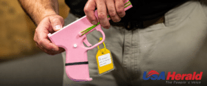 A person holds a pink plastic toy gun with a yellow tag attached, blurred background and USA Herald logo in the bottom right corner.