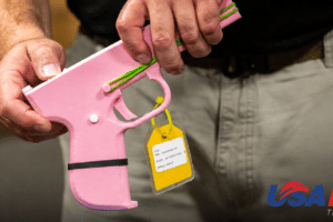A person holds a pink plastic toy gun with a yellow tag attached, blurred background and USA Herald logo in the bottom right corner.