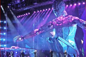 Geometric blue humanoid robot stage prop on a concert stage with purple lights and musicians performing.