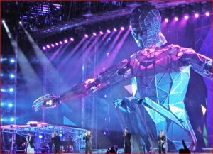 Geometric blue humanoid robot stage prop on a concert stage with purple lights and musicians performing.