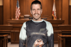 Smiling man stands in a courtroom with wooden panels and a flag behind him.