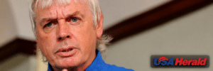 Close-up of a gray-haired man in a blue shirt speaking, with a blurred background and a USA Herald watermark in the bottom right corner.