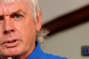 Close-up of a gray-haired man in a blue shirt speaking, with a blurred background and a USA Herald watermark in the bottom right corner.