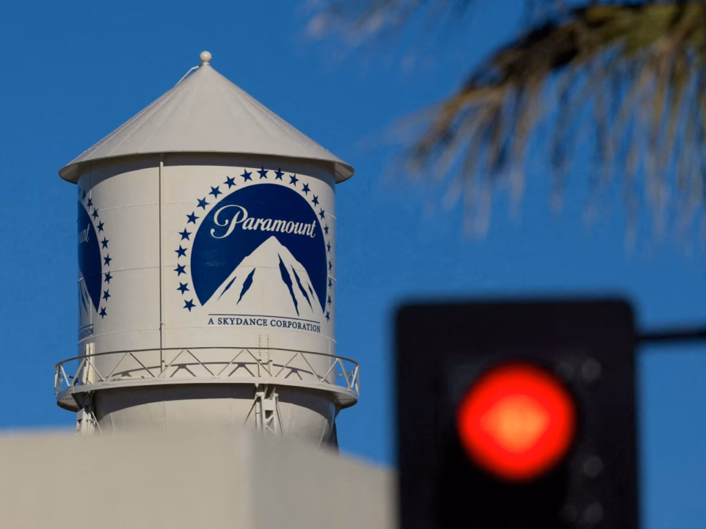 White Paramount water tower with a blue circular logo of mountains, set against a bright blue sky; a blurred red traffic light appears in the foreground.