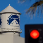 White Paramount water tower with a blue circular logo of mountains, set against a bright blue sky; a blurred red traffic light appears in the foreground.