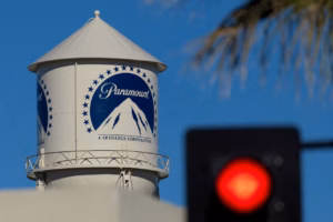 White Paramount water tower with a blue circular logo of mountains, set against a bright blue sky; a blurred red traffic light appears in the foreground.