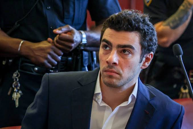 Man in a navy suit and white shirt sits in a courtroom, looking to the side with a tense expression; uniformed officers are in the background.