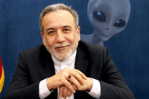 Man in a dark suit sits at a desk, hands folded, with the Iranian flag on his left and a gray alien figure in the background.