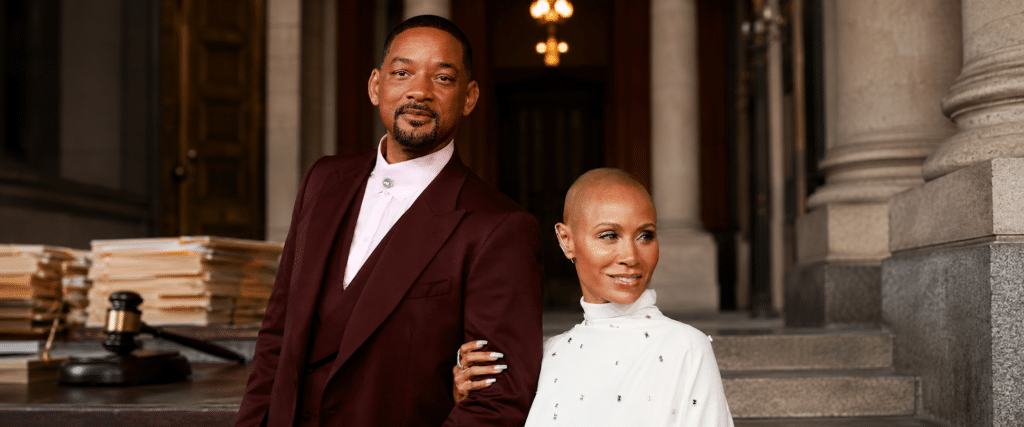 Two stylish adults pose arm-in-arm on stone steps outside a grand building, the man in a burgundy suit and the woman in white.