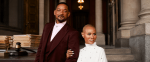Two stylish adults pose arm-in-arm on stone steps outside a grand building, the man in a burgundy suit and the woman in white.