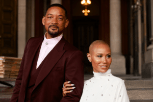 Two stylish adults pose arm-in-arm on stone steps outside a grand building, the man in a burgundy suit and the woman in white.