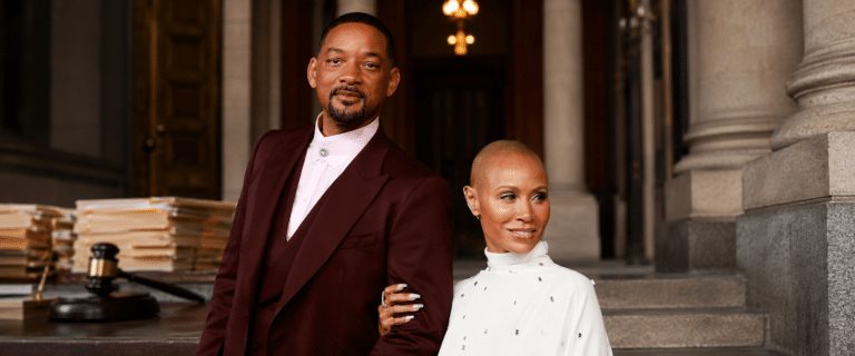 Two stylish adults pose arm-in-arm on stone steps outside a grand building, the man in a burgundy suit and the woman in white.