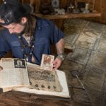 Bearded man in a blue shirt and cap reads an old newspaper at a wooden table in a rustic room.