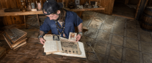 Bearded man in a blue shirt and cap reads an old newspaper at a wooden table in a rustic room.