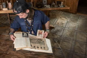 Bearded man in a blue shirt and cap reads an old newspaper at a wooden table in a rustic room.