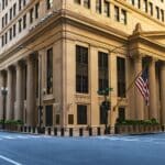 Beige neoclassical bank building with large columns at a street corner, an American flag hanging from the facade, and a crosswalk in the foreground at Jackson and La Salle streets.
