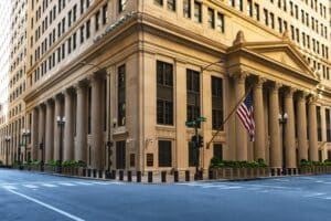 Beige neoclassical bank building with large columns at a street corner, an American flag hanging from the facade, and a crosswalk in the foreground at Jackson and La Salle streets.