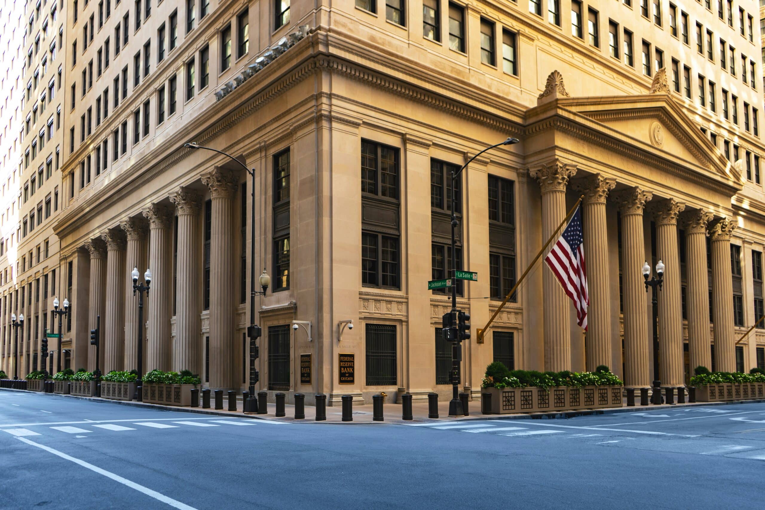 Beige neoclassical bank building with large columns at a street corner, an American flag hanging from the facade, and a crosswalk in the foreground at Jackson and La Salle streets.
