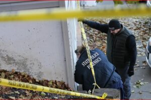 Forensic investigators at a crime scene outside a house, yellow police tape in the foreground.
