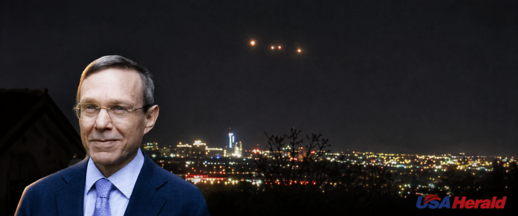 Portrait of a man in a blue suit with a night city skyline behind him; USA Herald watermark in the bottom right corner.