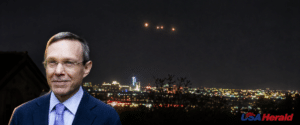 Portrait of a man in a blue suit with a night city skyline behind him; USA Herald watermark in the bottom right corner.
