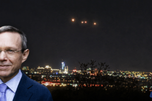 Portrait of a man in a blue suit with a night city skyline behind him; USA Herald watermark in the bottom right corner.