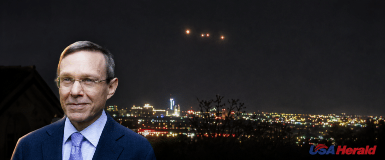 Portrait of a man in a blue suit with a night city skyline behind him; USA Herald watermark in the bottom right corner.