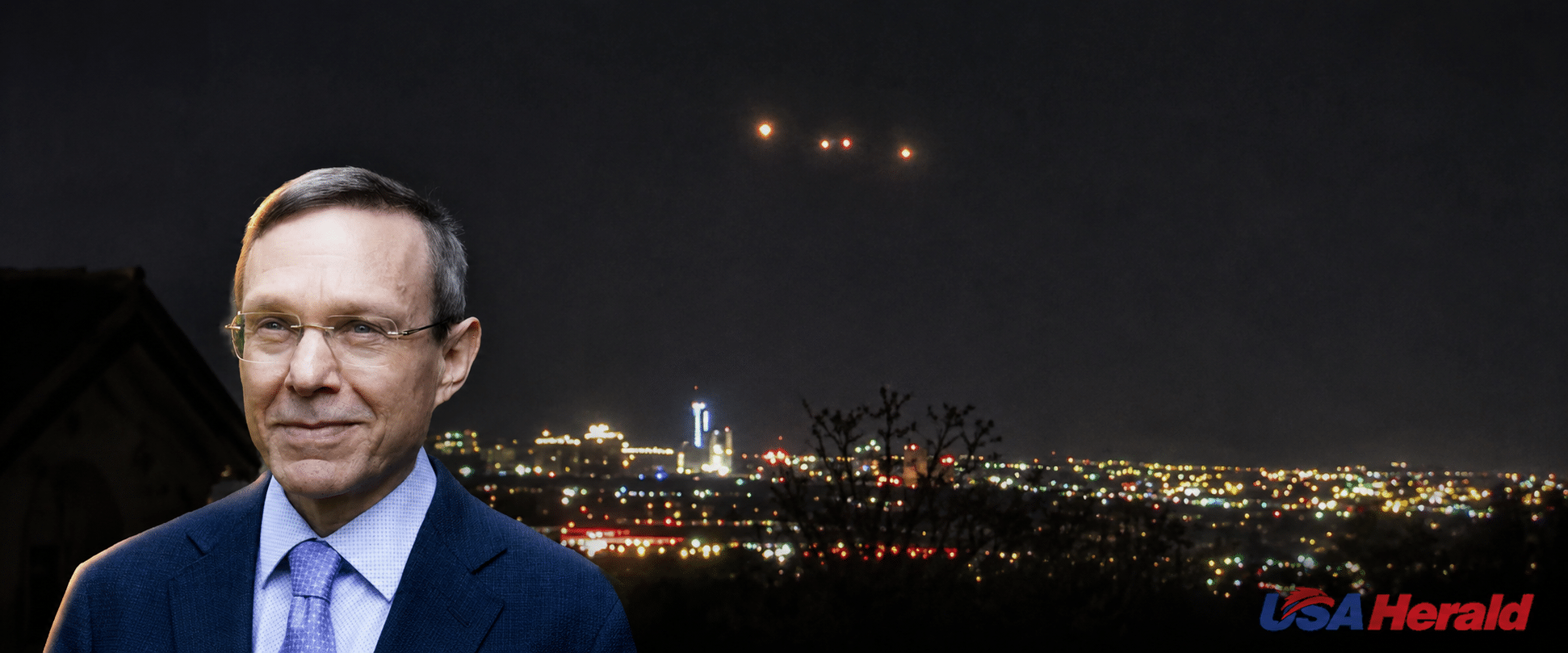 Portrait of a man in a blue suit with a night city skyline behind him; USA Herald watermark in the bottom right corner.