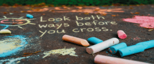 Pavement chalk message reading 'Look both ways before you cross' with pastel chalk pieces nearby.