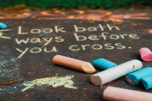 Pavement chalk message reading 'Look both ways before you cross' with pastel chalk pieces nearby.