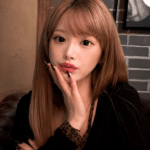Portrait of a young woman with long light brown hair sitting on a leather sofa in a dim bar, touching her lips with her hand and black nails visible, bottles on a shelf in the background.