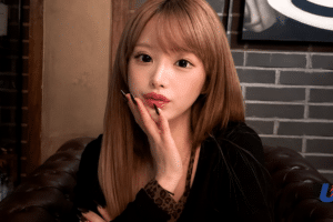 Portrait of a young woman with long light brown hair sitting on a leather sofa in a dim bar, touching her lips with her hand and black nails visible, bottles on a shelf in the background.