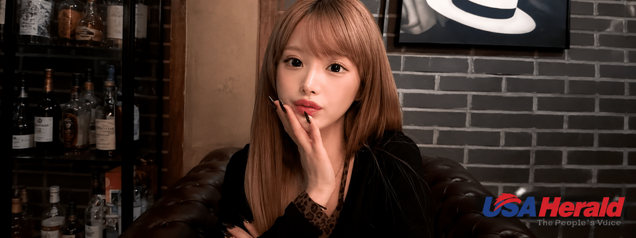 Portrait of a young woman with long light brown hair sitting on a leather sofa in a dim bar, touching her lips with her hand and black nails visible, bottles on a shelf in the background.