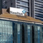 Steadfast sign on a modern glass office building with a tan roof, a tall dark high-rise behind it.