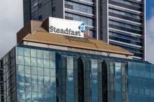 Steadfast sign on a modern glass office building with a tan roof, a tall dark high-rise behind it.