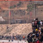 Large crowd of people along a rocky seawall next to a tall fence and security posts in the distance.