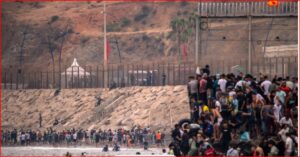 Large crowd of people along a rocky seawall next to a tall fence and security posts in the distance.