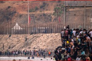 Large crowd of people along a rocky seawall next to a tall fence and security posts in the distance.