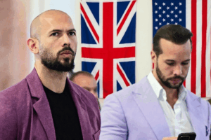 Two men in blazers stand indoors with British and American flags in the background, looking ahead (one with a shaved head and beard).