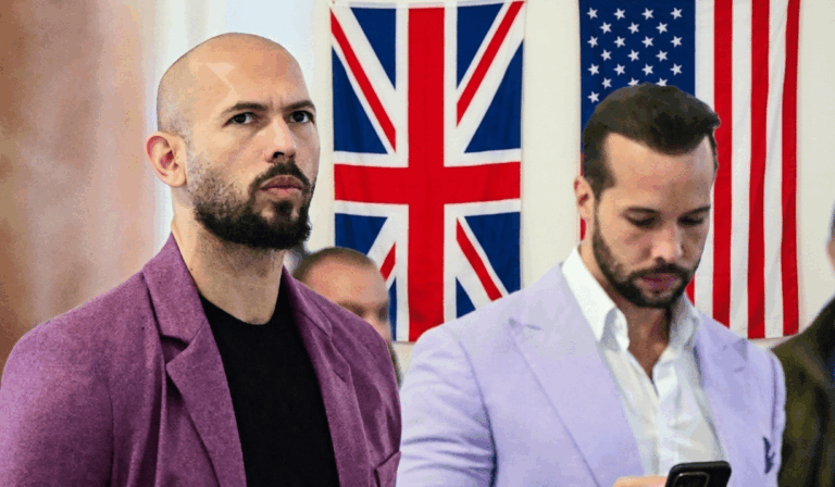 Two men in blazers stand indoors with British and American flags in the background, looking ahead (one with a shaved head and beard).