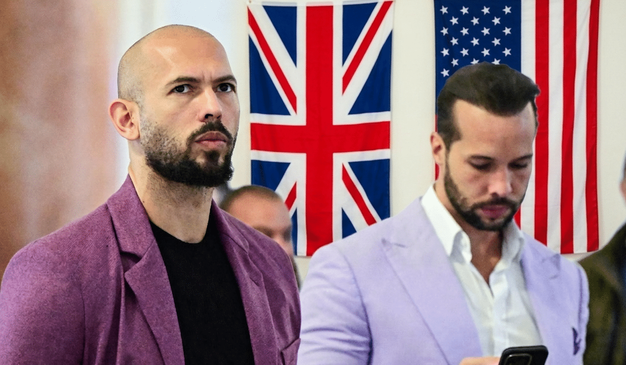 Two men in blazers stand indoors with British and American flags in the background, looking ahead (one with a shaved head and beard).