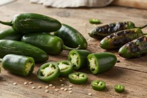 Fresh and roasted jalapeño peppers on a wooden board, some sliced open to reveal seeds and pale interior of the peppers.