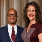 Two professionals smiling side by side in a law library, with a gavel and books nearby. The man wears a navy suit and red polka-dot tie, and the woman wears a burgundy dress with a checked pattern.