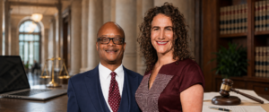 Two professionals smiling side by side in a law library, with a gavel and books nearby. The man wears a navy suit and red polka-dot tie, and the woman wears a burgundy dress with a checked pattern.