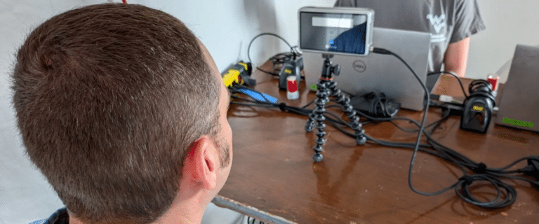 Back of a man’s head at a desk, smartphone on a small tripod among cables and tech gear.