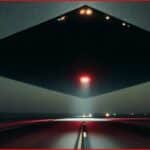 Gigantic, dark, square UFO hovering over a desert highway at night with a red central glow.